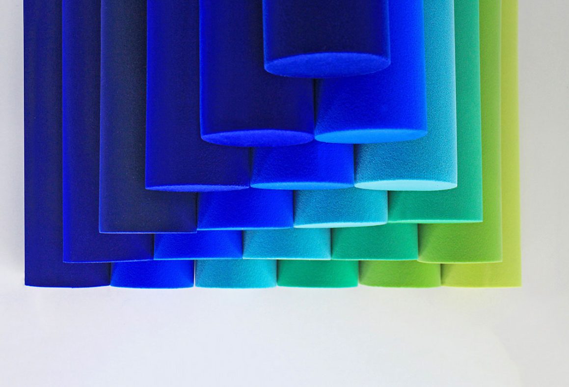 Rows of cylindrical foam tubes in gradient shades from deep blue to green are arranged in a neat pattern against a white background. The tubes overlap, creating a visually appealing transition of colors.