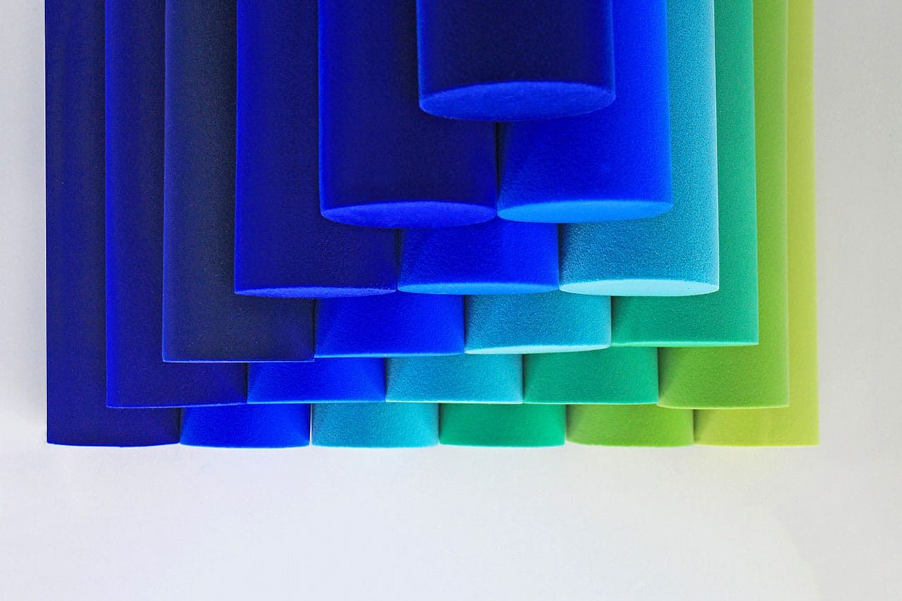 Rows of cylindrical foam tubes in gradient shades from deep blue to green are arranged in a neat pattern against a white background. The tubes overlap, creating a visually appealing transition of colors.