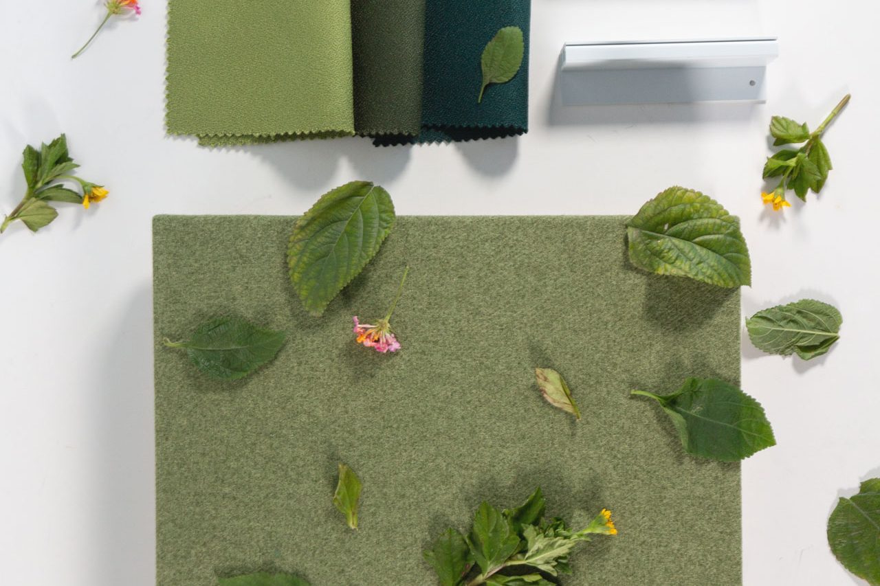 Three fabric swatches in shades of green and teal, three gray rectangular objects, and a green textured square are arranged on a white surface, with scattered leaves and small colorful flowers.