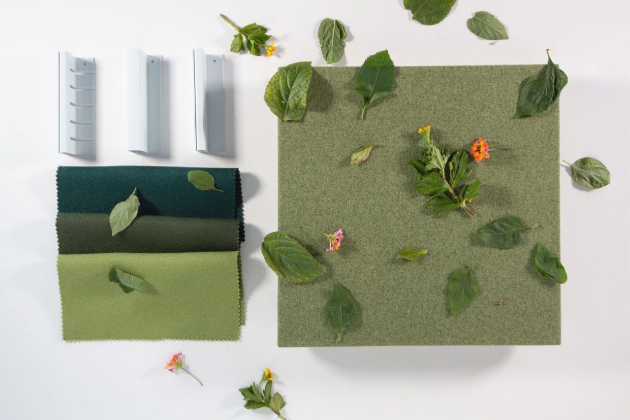 A green fabric swatch, several green leaves, small flowers, and three gray plastic objects are arranged neatly on a white surface. The green tones vary from dark to light.