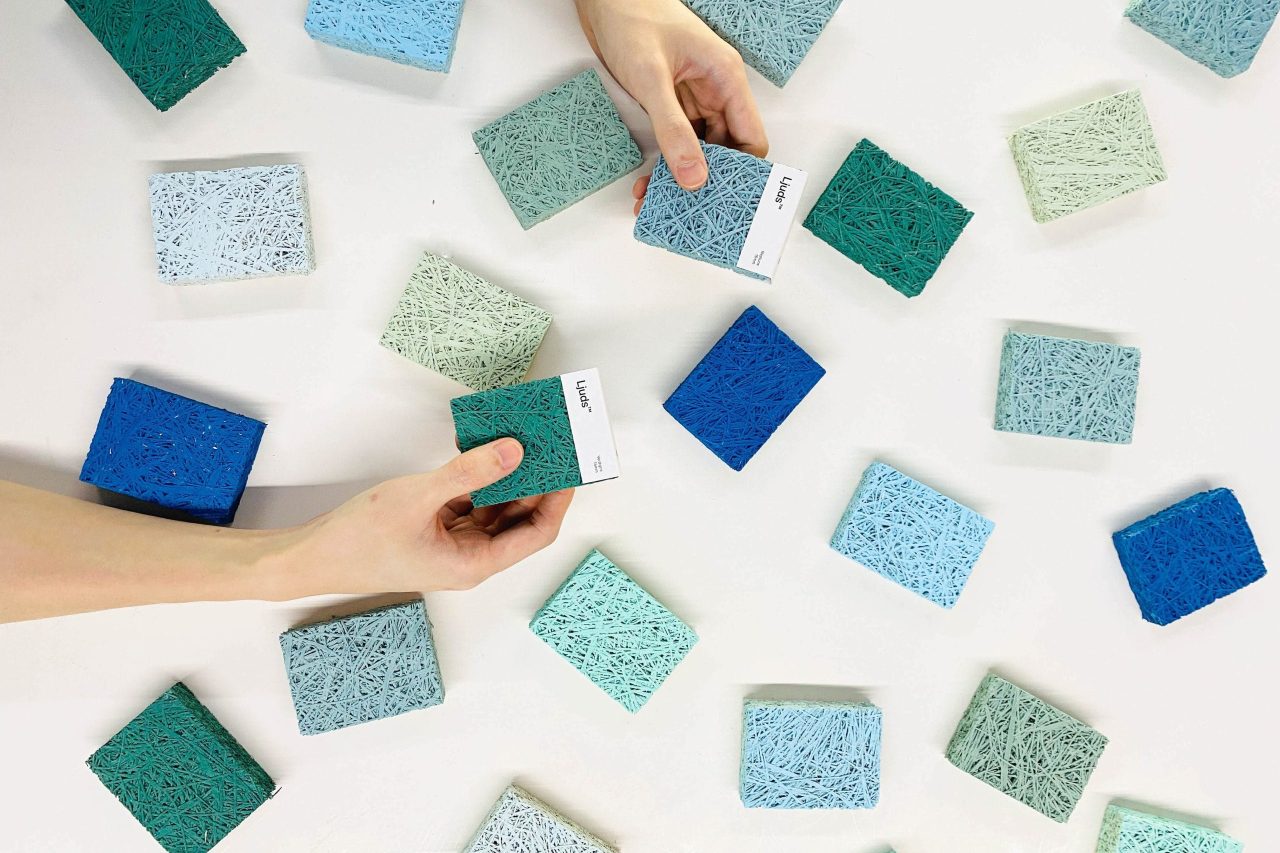 Two hands hold textured, rectangular color samples above a surface scattered with similar swatches in shades of blue, teal, and light green. The samples have a fibrous, woven appearance.