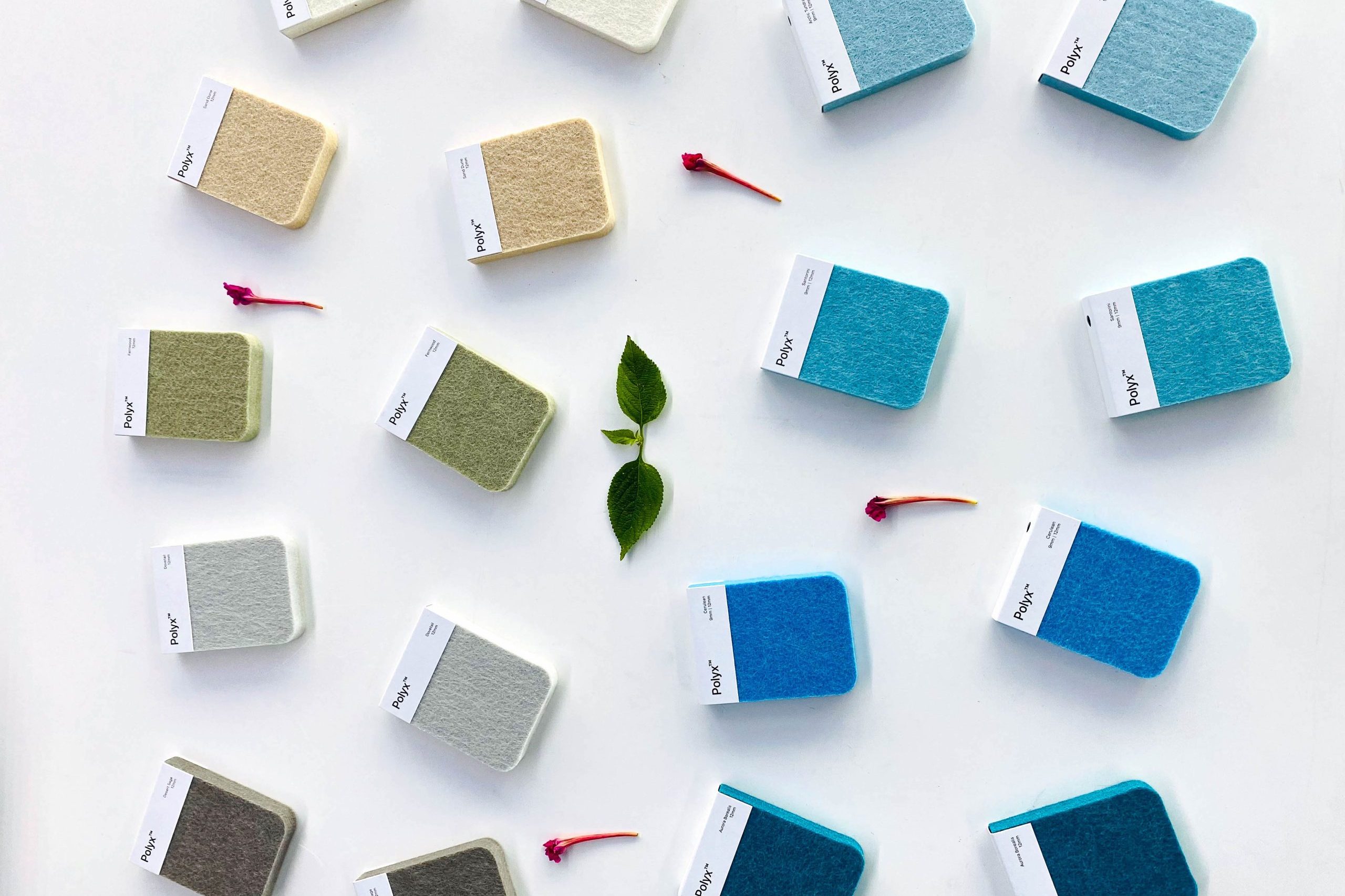 Fabric swatches in shades of blue, green, gray, and beige are arranged in a circular pattern on a white surface, with three green leaves and small red flower stems in the center.
