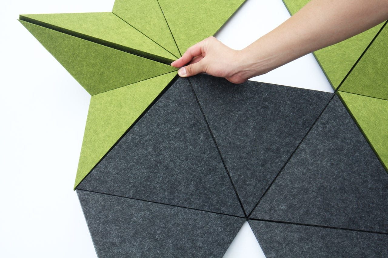 A hand arranges triangular, felt-like tiles in shades of green and dark gray on a white surface, forming a geometric, star-shaped pattern.