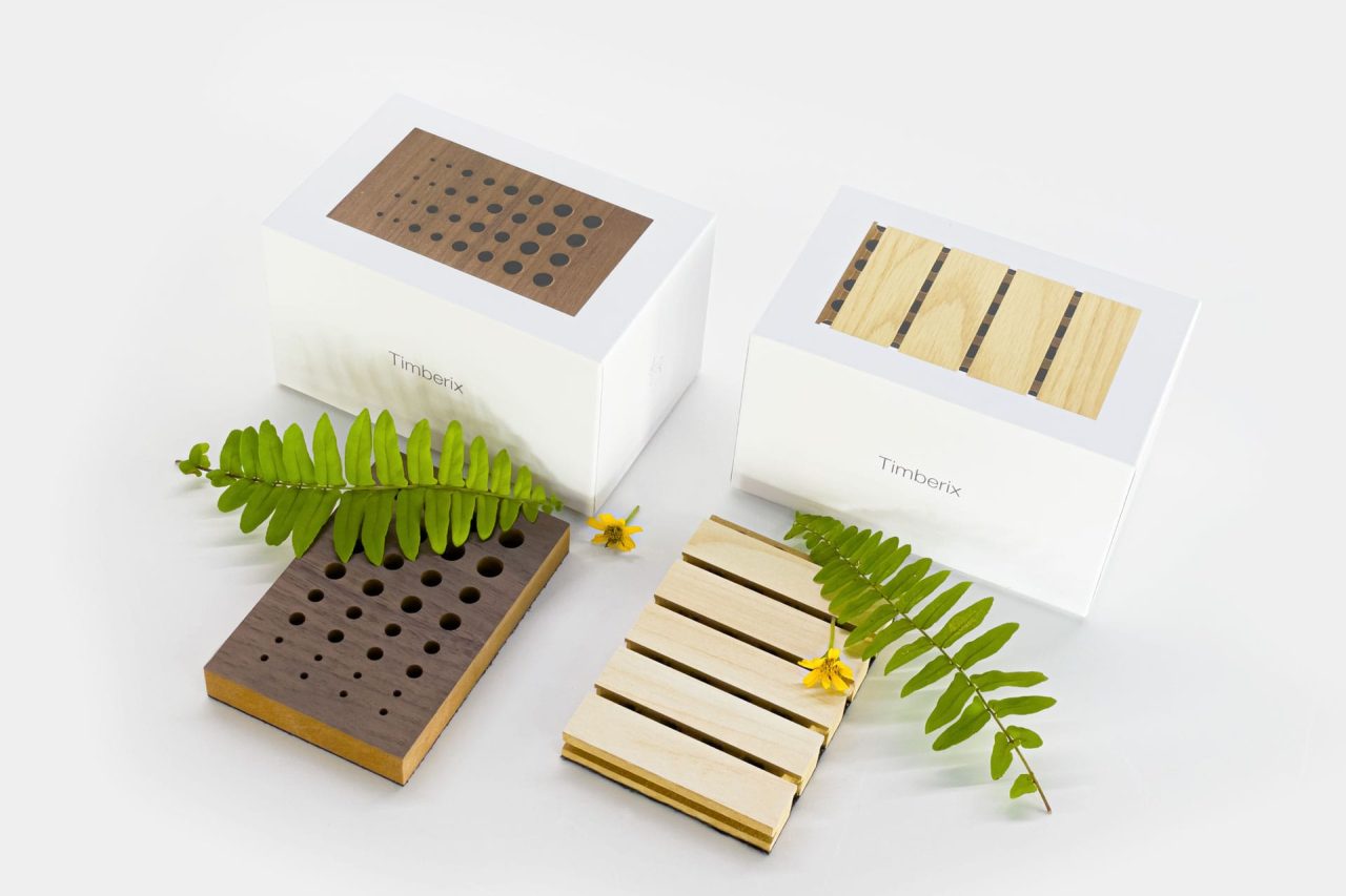 Two white boxes with wooden tops, one featuring round holes and the other parallel slats, are placed on a white surface with green fern leaves and yellow flowers for decoration.